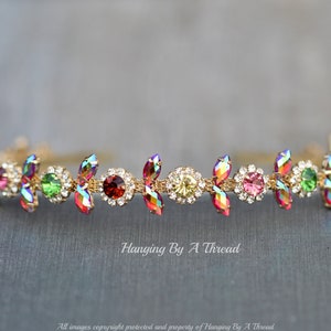 May include: A gold headband with iridescent red, green, and clear rhinestones. The headband is decorated with small, gold leaves.