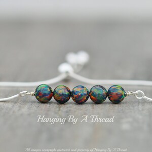 May include: A silver chain bracelet with five iridescent black beads and a sliding closure. The bracelet is on a wooden surface. "Hanging By A Thread" is written below the bracelet.