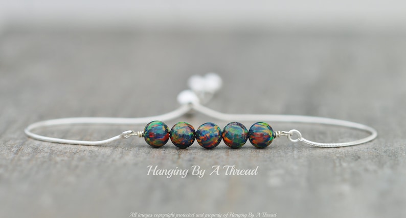 May include: A silver chain bracelet with five iridescent black beads and a sliding closure. The bracelet is on a wooden surface. "Hanging By A Thread" is written below the bracelet.