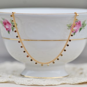 Puede incluir: Un collar de cadena dorada con peque&ntilde;os colgantes redondos. El collar se muestra en una taza de t&eacute; de porcelana blanca.