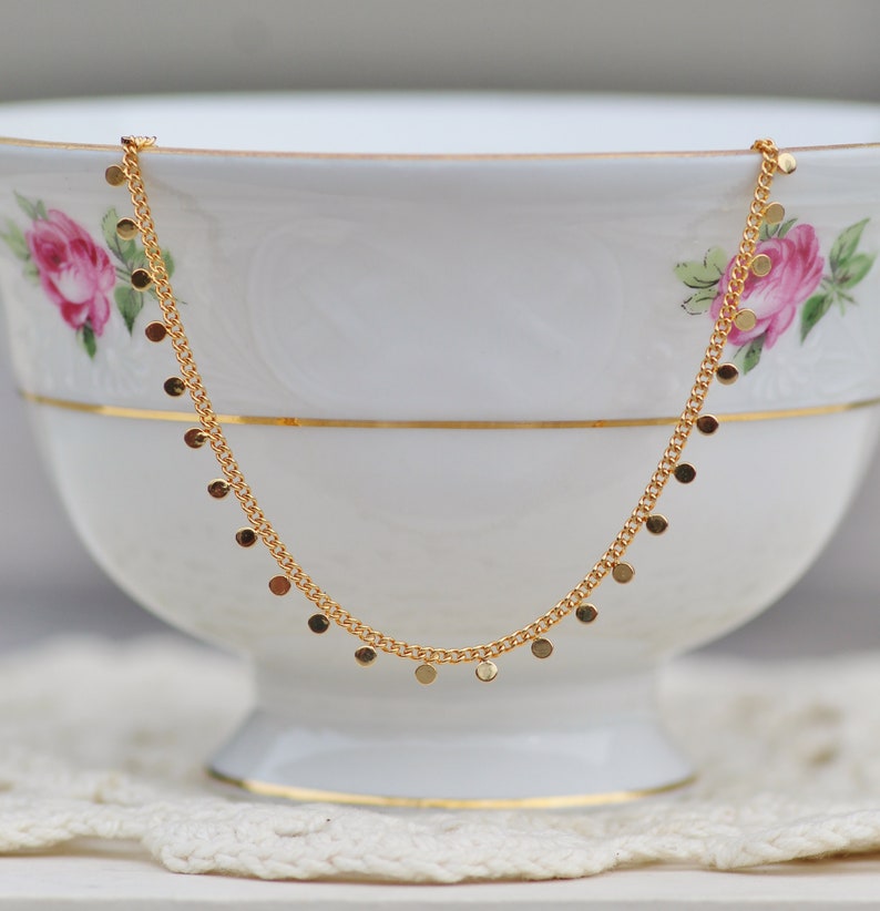 Puede incluir: Un collar de cadena dorada con peque&ntilde;os colgantes redondos. El collar se muestra en una taza de t&eacute; de porcelana blanca.