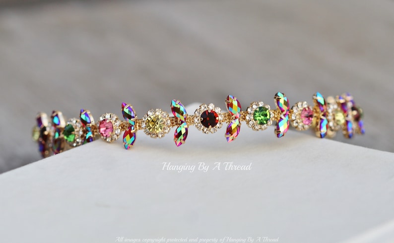 May include: A gold headband with a row of iridescent, multi-colored crystals in a floral design. The crystals are set in a gold metal frame and are arranged in a repeating pattern. The headband is a delicate and elegant accessory.