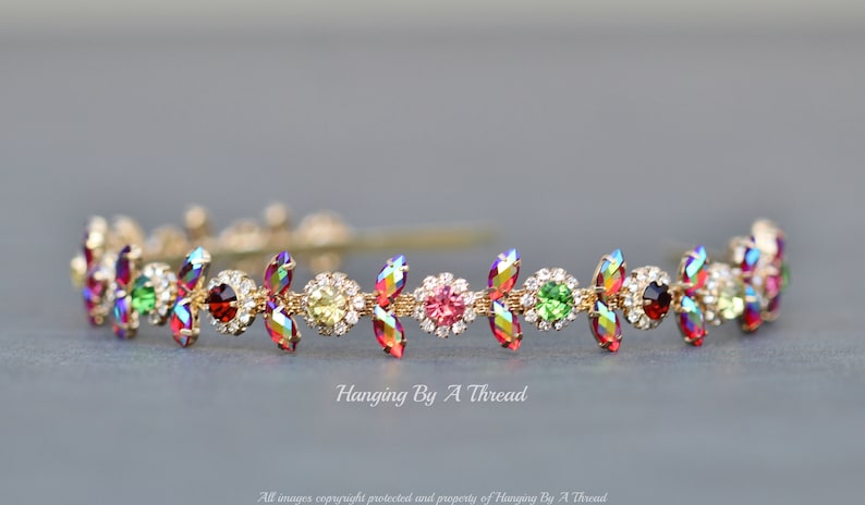 May include: A gold headband with a row of colorful rhinestones in a floral design. The rhinestones are red, green, pink, and clear.