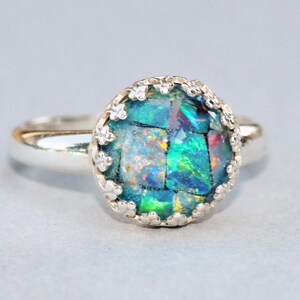 May include: A silver ring with a mosaic-style, iridescent gemstone. The gemstone is primarily teal and green, with flashes of other colors. The ring has a decorative, scalloped bezel setting.