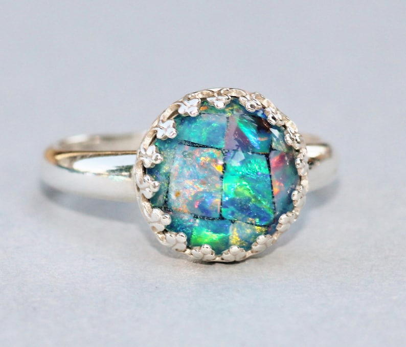 May include: A silver ring with a mosaic-style, iridescent gemstone. The gemstone is primarily teal and green, with flashes of other colors. The ring has a decorative, scalloped bezel setting.