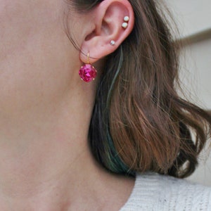 May include: A pair of pink gemstone earrings with gold hooks. The earrings are worn by a person with brown hair and a white sweater.