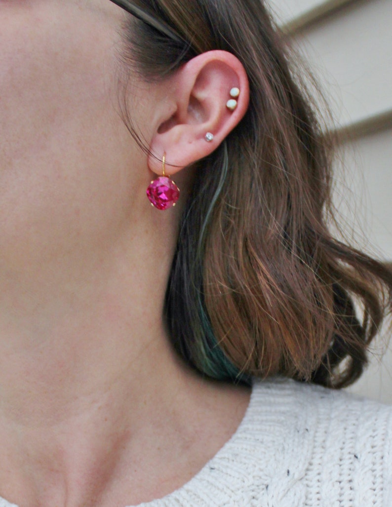 May include: A pair of pink gemstone earrings with gold hooks. The earrings are worn by a person with brown hair and a white sweater.