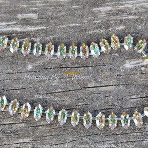 May include: A strand of iridescent, marquise-shaped rhinestones on a clear backing. The crystals reflect a rainbow of colors, including gold, green, and blue. The background is a weathered, gray wooden surface. Text on the image reads "Hanging By A Thread".