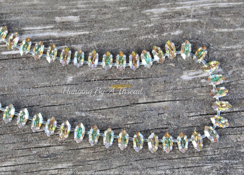 May include: A strand of iridescent, marquise-shaped rhinestones on a clear backing. The crystals reflect a rainbow of colors, including gold, green, and blue. The background is a weathered, gray wooden surface. Text on the image reads "Hanging By A Thread".
