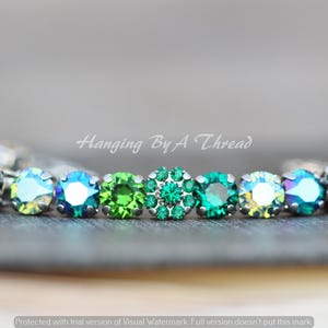 Puede incluir: Una pulsera con una fila de piedras preciosas verdes y verde azulado. Las piedras son redondas y están engastadas en un metal plateado. El texto "Hanging By A Thread" se ve en el fondo.