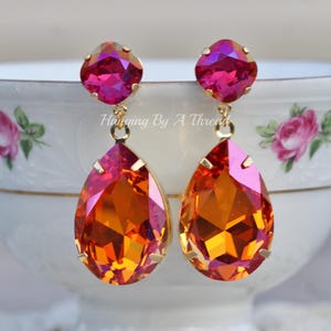 May include: A pair of dangle earrings featuring a teardrop-shaped orange and pink crystal, with a smaller round pink crystal at the top. The earrings are gold-toned.