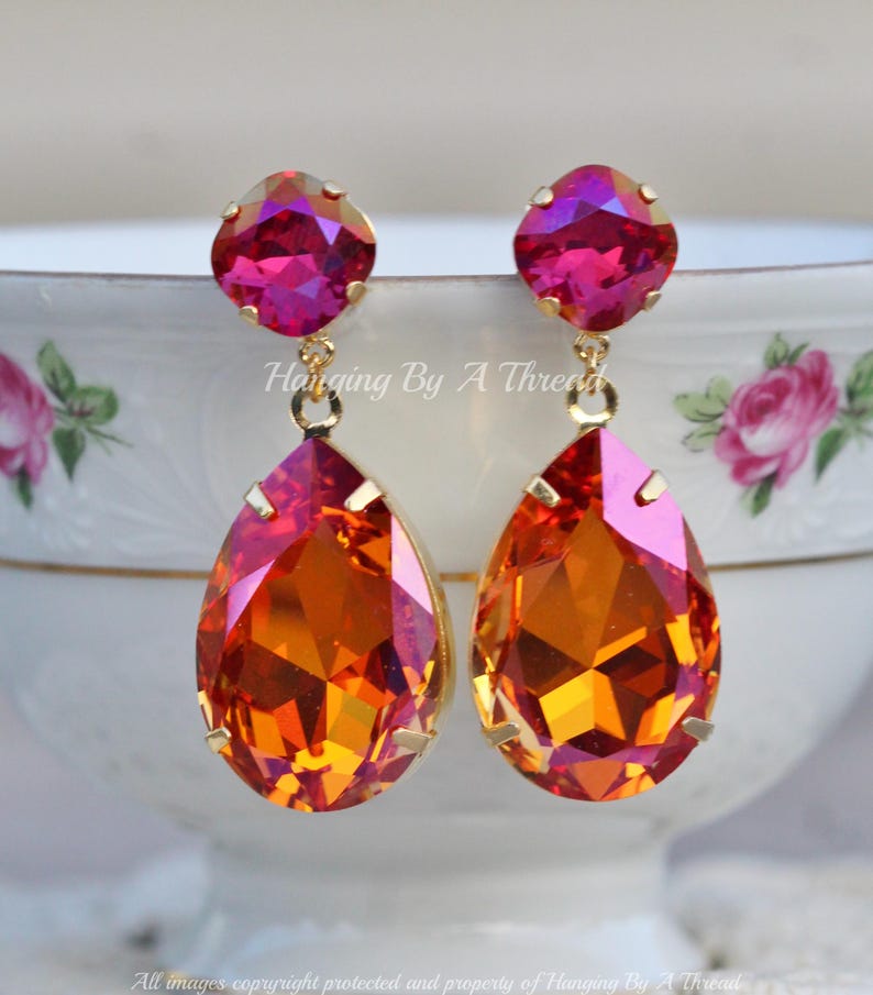 May include: A pair of dangle earrings featuring a teardrop-shaped orange and pink crystal, with a smaller round pink crystal at the top. The earrings are gold-toned.