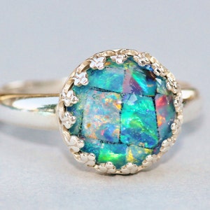 May include: A silver ring featuring a large, round, mosaic-style opal gemstone. The opal displays vibrant colors of green, blue, and hints of red. The setting has a delicate, floral-like design around the gemstone.