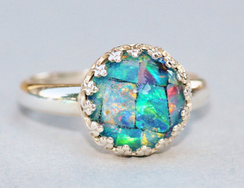 May include: A silver ring featuring a large, round, mosaic-style opal gemstone. The opal displays vibrant colors of green, blue, and hints of red. The setting has a delicate, floral-like design around the gemstone.