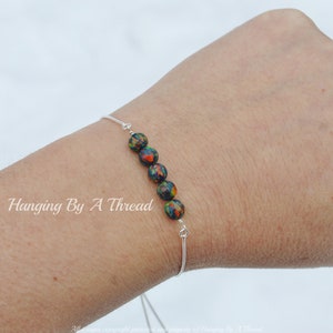 May include: A silver bracelet with a row of black, green, and orange beads. The bracelet is on a person's wrist. The text "Hanging By A Thread" is visible on the wrist.