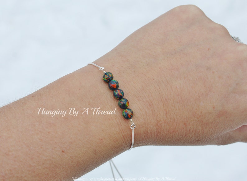 May include: A silver bracelet with a row of black, green, and orange beads. The bracelet is on a person's wrist. The text "Hanging By A Thread" is visible on the wrist.