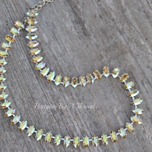 May include: A delicate necklace featuring iridescent, marquise-shaped crystals set in a silver-tone chain. The crystals shift in color, displaying hues of yellow, green, and gold. The necklace is displayed on a wooden surface. Text on the image reads "Hanging By A Thread".