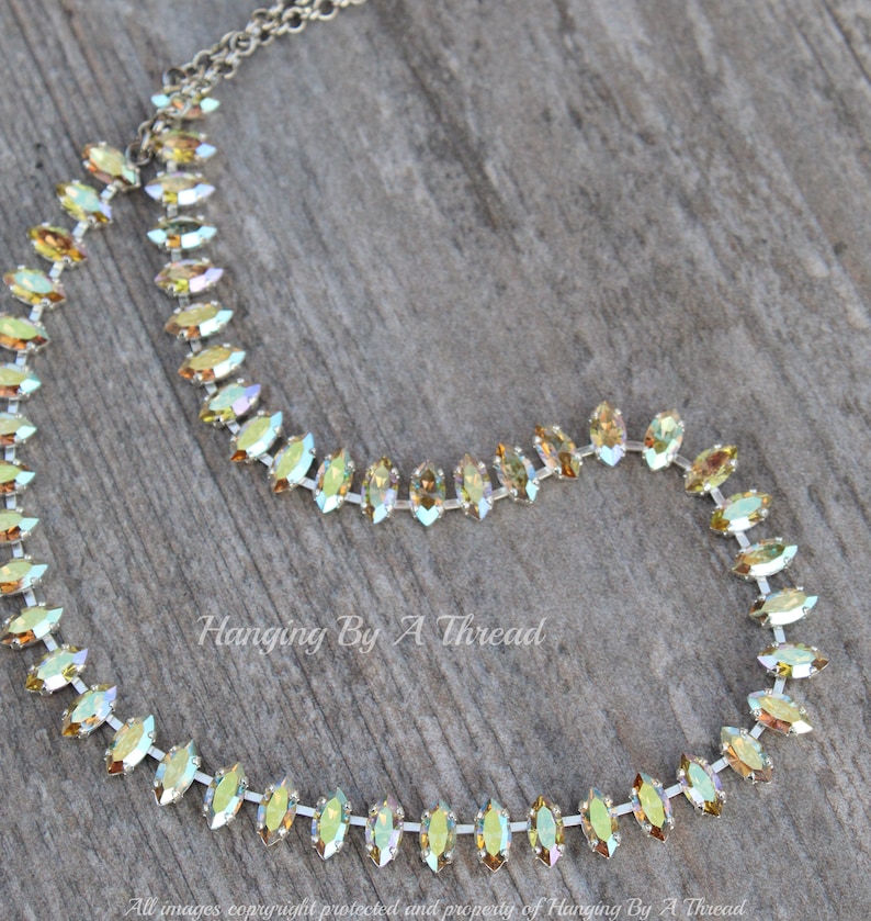 May include: A delicate necklace featuring iridescent, marquise-shaped crystals set in a silver-tone chain. The crystals shift in color, displaying hues of yellow, green, and gold. The necklace is displayed on a wooden surface. Text on the image reads "Hanging By A Thread".