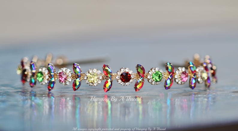 May include: A gold headband with a row of colorful rhinestones. The rhinestones are in a variety of shapes and sizes, including round, oval, and marquise. The headband is a delicate and elegant accessory.