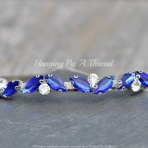 Pu&ograve; includere: Un cerchietto argentato con una fila di strass blu e trasparenti a forma di marquise. Il cerchietto &egrave; curvo e presenta un design delicato. Hanging By A Thread.