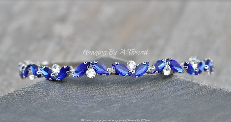 Pu&ograve; includere: Un cerchietto argentato con una fila di strass blu e trasparenti a forma di marquise. Il cerchietto &egrave; curvo e presenta un design delicato. Hanging By A Thread.