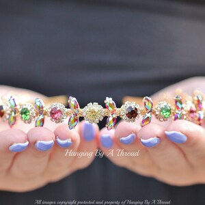 May include: A gold headband with iridescent, multi-colored gemstones. The headband is decorated with small, clear rhinestones.