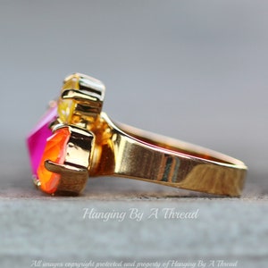 May include: A gold-toned ring featuring a cluster of colorful gemstones. The ring showcases a large, faceted, hot pink gemstone, complemented by orange and yellow stones. The band is smooth and polished, with the text "Hanging By A Thread" visible.