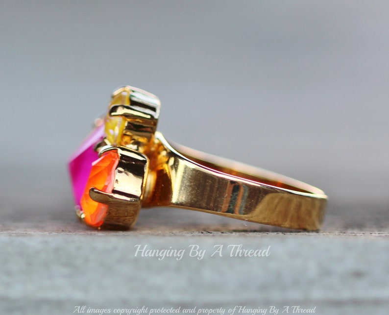 May include: A gold-toned ring featuring a cluster of colorful gemstones. The ring showcases a large, faceted, hot pink gemstone, complemented by orange and yellow stones. The band is smooth and polished, with the text "Hanging By A Thread" visible.