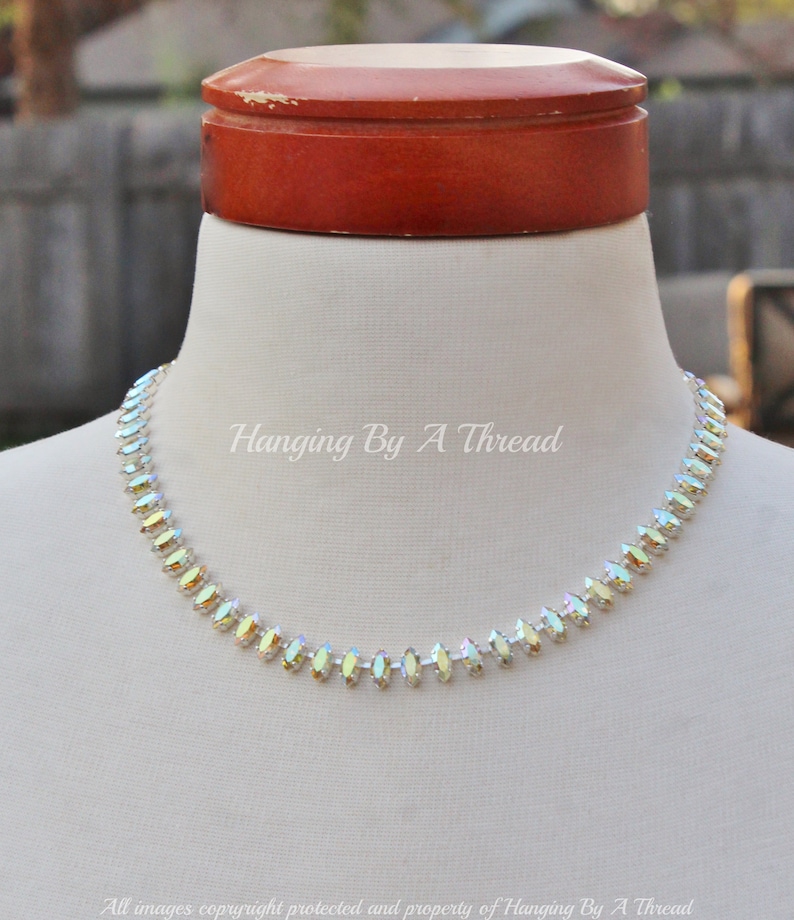 May include: A delicate necklace with iridescent, marquise-shaped crystals. The crystals are set in a row, creating a sparkling choker style. The text "Hanging By A Thread" is visible on the image.
