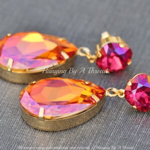 May include: A pair of gold dangle earrings featuring a teardrop-shaped orange and pink iridescent crystal, and a smaller pink crystal. The earrings are labelled "Hanging By A Thread".