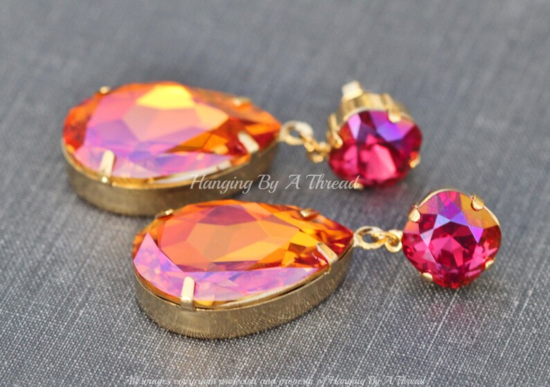 May include: A pair of gold dangle earrings featuring a teardrop-shaped orange and pink iridescent crystal, and a smaller pink crystal. The earrings are labelled "Hanging By A Thread".