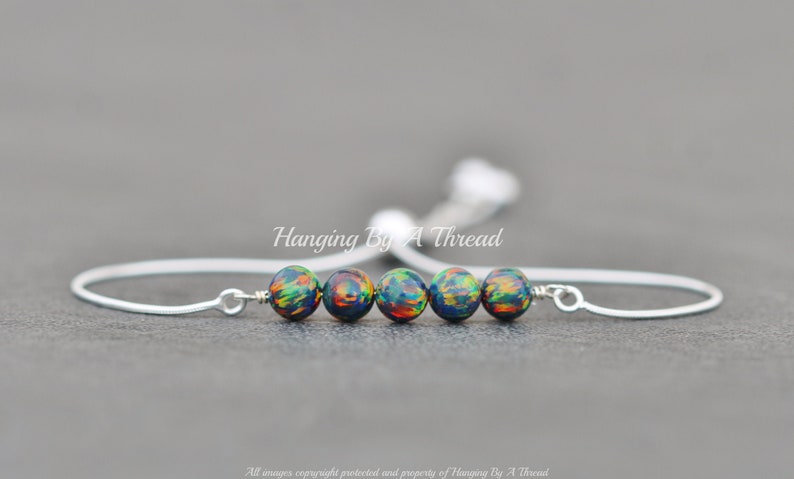 May include: A silver adjustable bracelet with a row of five black, green, and orange opal beads. The bracelet is tied with a silver cord and has the text "Hanging By A Thread" on the cord.