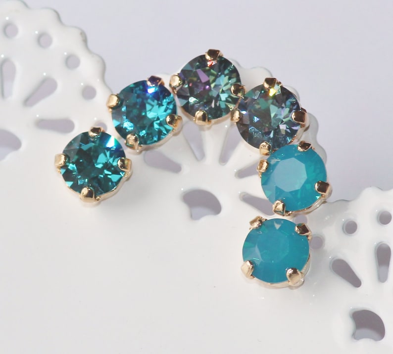 May include: A set of six turquoise and teal blue rhinestone earrings with gold settings. The rhinestones are round and have a faceted finish.