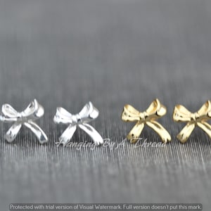 Pode incluir: Quatro pequenos brincos em forma de laço, dois prateados e dois dourados, expostos em uma superfície cinza.