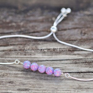 Puede incluir: Un brazalete ajustable de plata con una fila de seis cuentas iridiscentes de color p&uacute;rpura y rosa. El brazalete est&aacute; sobre una superficie de madera.