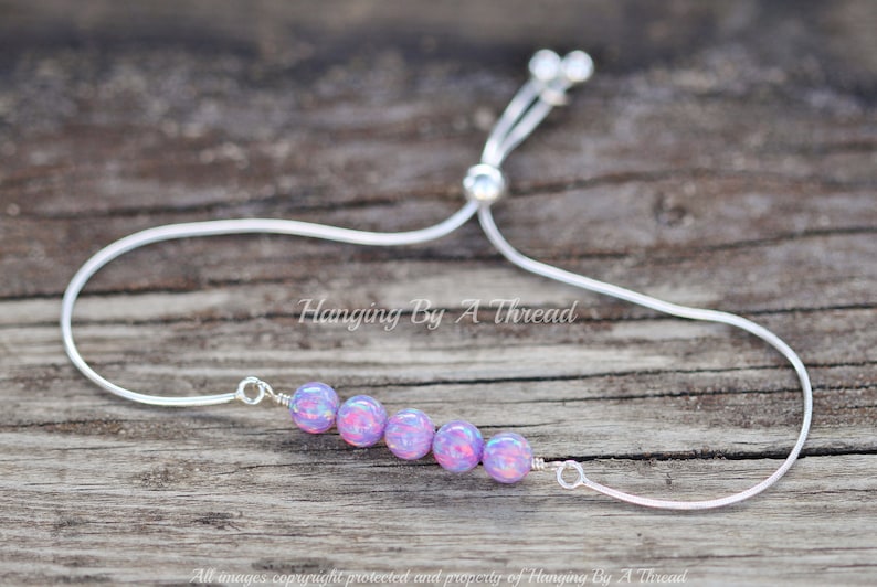Puede incluir: Un brazalete ajustable de plata con una fila de seis cuentas iridiscentes de color p&uacute;rpura y rosa. El brazalete est&aacute; sobre una superficie de madera.