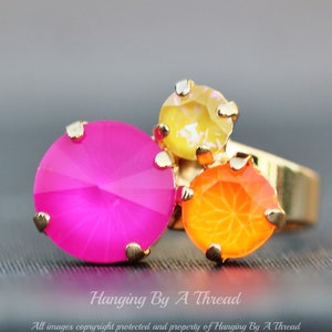 May include: A gold-toned ring featuring three colorful, faceted gemstones. The largest is a vibrant pink, flanked by an orange and a yellow stone. The ring is set against a blurred gray background.