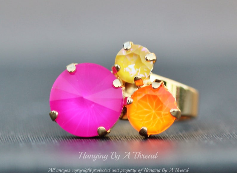 May include: A gold-toned ring featuring three colorful, faceted gemstones. The largest is a vibrant pink, flanked by an orange and a yellow stone. The ring is set against a blurred gray background.