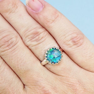 May include: A silver ring with a large, round, iridescent blue-green stone. The stone is set in a decorative, scalloped bezel. The ring is shown on a person's finger against a light blue background.