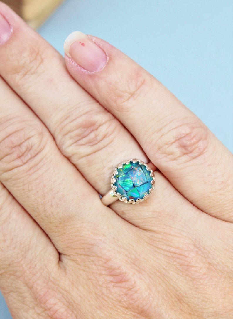 May include: A silver ring with a large, round, iridescent blue-green stone. The stone is set in a decorative, scalloped bezel. The ring is shown on a person's finger against a light blue background.