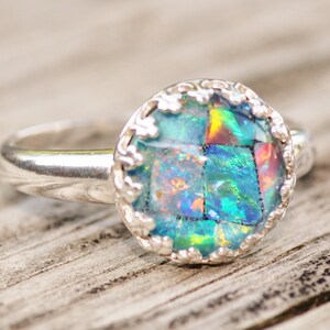 May include: A silver ring with a round, mosaic-style opal stone. The opal features a vibrant display of blues, greens, and hints of red. The ring's setting has a decorative, scalloped edge, adding a touch of elegance to the jewelry.