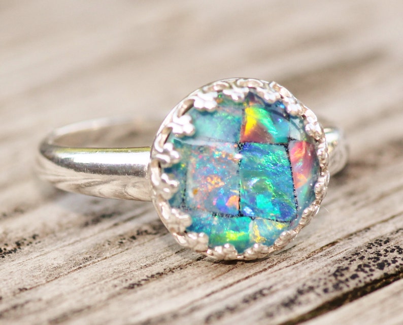 May include: A silver ring with a round, mosaic-style opal stone. The opal features a vibrant display of blues, greens, and hints of red. The ring's setting has a decorative, scalloped edge, adding a touch of elegance to the jewelry.