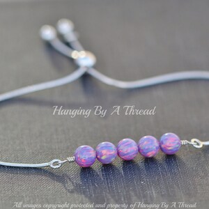 Puede incluir: Un brazalete ajustable de plata con una fila de seis cuentas iridiscentes de color p&uacute;rpura y rosa. El brazalete est&aacute; atado con un cord&oacute;n de plata y tiene un cierre de plata.