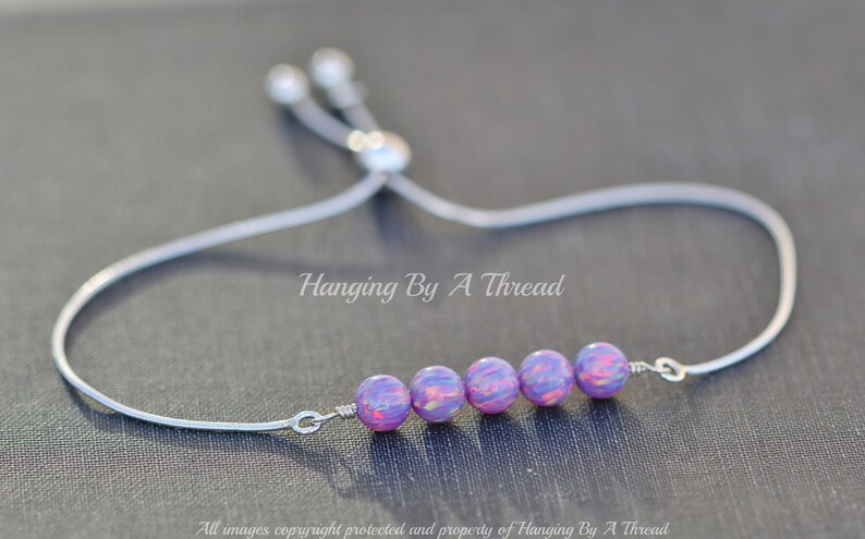 Puede incluir: Un brazalete ajustable de plata con una fila de seis cuentas iridiscentes de color p&uacute;rpura y rosa. El brazalete est&aacute; atado con un cord&oacute;n de plata y tiene un cierre de plata.