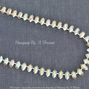 May include: A strand of iridescent rhinestones in a marquise shape, strung on a white backing. The rhinestones have a shimmering, multi-colored effect. The text "Hanging By A Thread" is visible in the image.