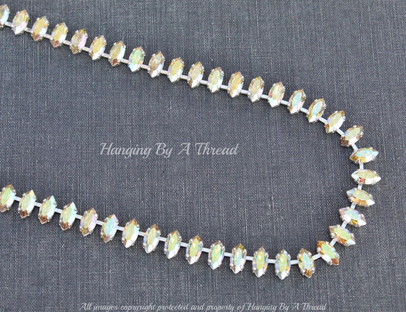 May include: A strand of iridescent rhinestones in a marquise shape, strung on a white backing. The rhinestones have a shimmering, multi-colored effect. The text "Hanging By A Thread" is visible in the image.