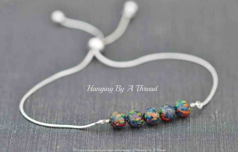 May include: A silver chain bracelet with a sliding knot closure. The bracelet features five round, dark blue beads with colorful flecks. The text "Hanging By A Thread" is visible below the bracelet.