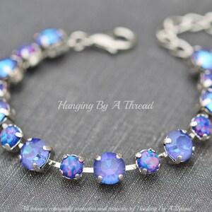 May include: A silver chain bracelet with a row of iridescent blue and purple round crystals. The bracelet is on a gray fabric background. Hanging By A Thread.