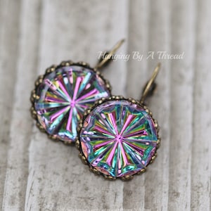 Puede incluir: Un par de pendientes de cristal iridiscente con un diseño de estrella, engastados en monturas de latón antiguo. Los pendientes cuelgan de una superficie de madera.