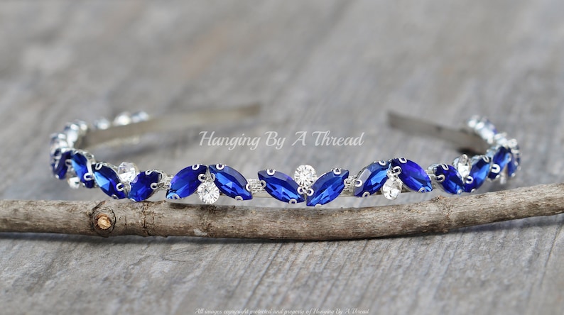 Pu&ograve; includere: Un cerchietto argentato con una fila di strass blu e trasparenti. Il cerchietto &egrave; su una superficie di legno. Hanging By A Thread.